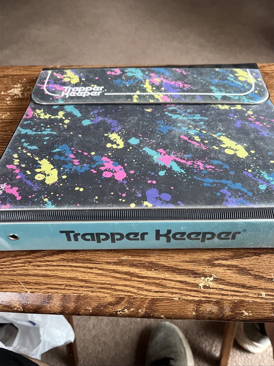 A colorful Trapper Keeper binder with splatter paint design sits on a wooden table. The binder has a Velcro flap and the words "Trapper Keeper" printed on the spine and cover.