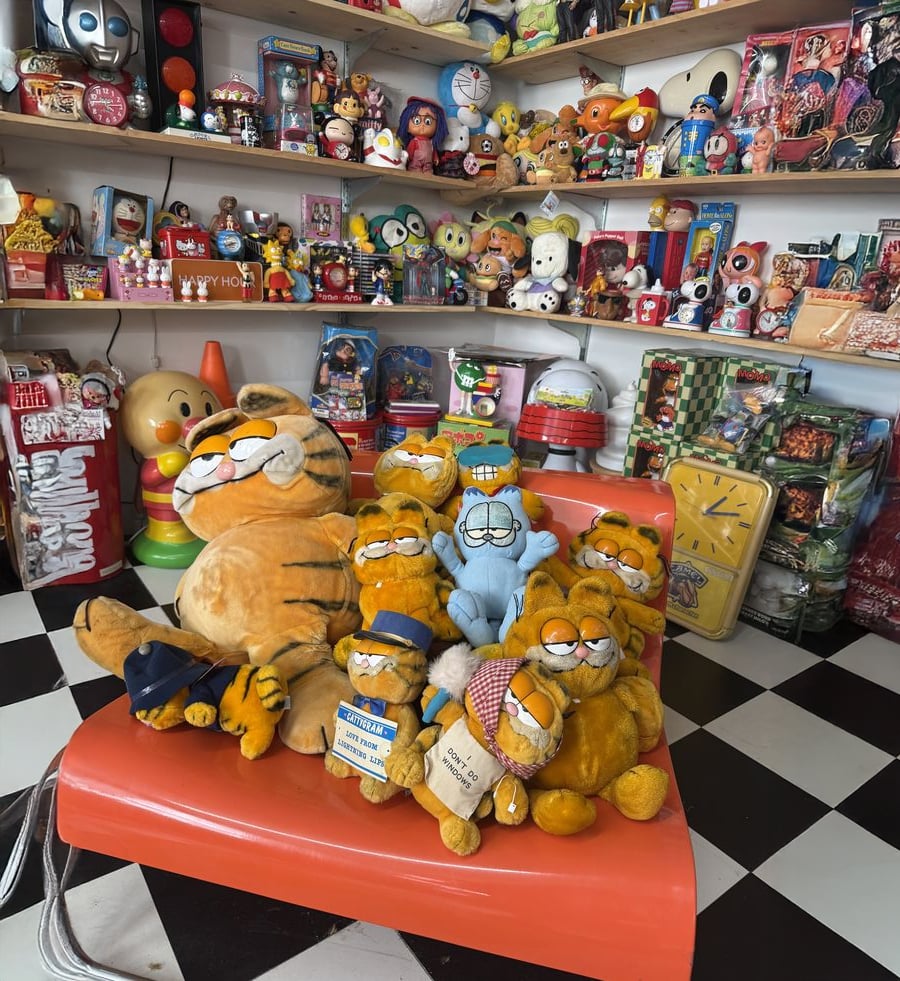 A group of Garfield plush toys sit on an orange bench in a store filled with colorful vintage toys and collectibles displayed on shelves against a checkered black-and-white floor.