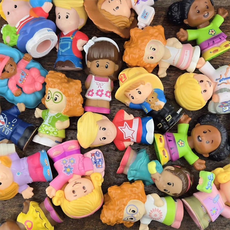 A variety of small, colorful plastic toy figures with different outfits and hairstyles are scattered on a wooden surface. Most figures are lying on their sides or backs, facing different directions.