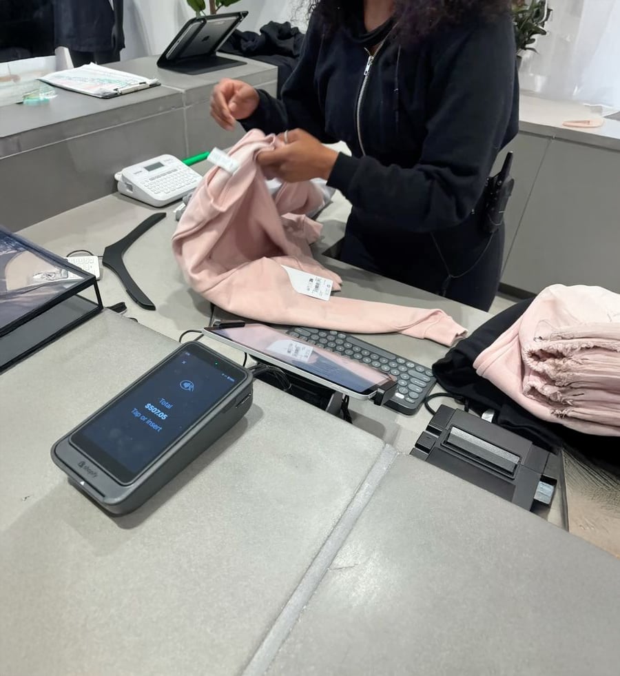 A retail worker folds pink clothing behind a checkout counter with a payment terminal displaying a digital wallet ready for contactless payment. Other folded clothes and store equipment are visible on the counter.