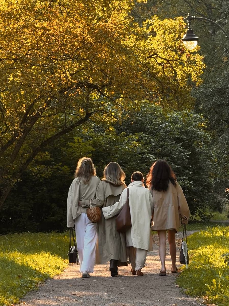 Four women walk arm in arm down a sunlit path lined with trees, dressed in neutral-toned coats and carrying bags, surrounded by lush greenery and golden evening light.