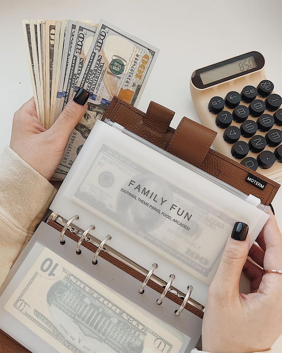 A person holds a stack of U.S. dollar bills in one hand and a brown budget binder labeled "Family Fun" in the other, with a calculator displaying 95.1 in the background.