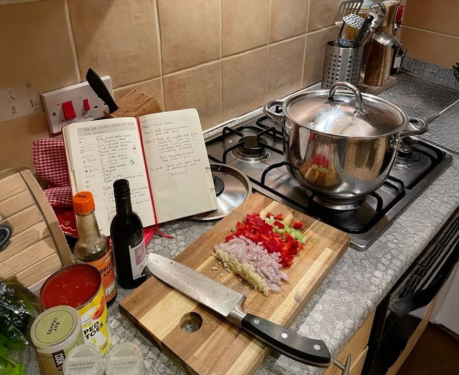 A kitchen countertop with a chopping board holding chopped shallots, garlic, ginger, and red pepper, a large knife, a notebook with handwritten recipes, various condiments, and a pot on the stove.