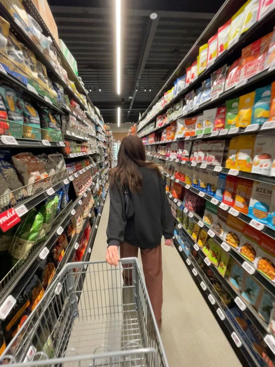 A person with long brown hair, wearing a black jacket and brown pants, pushes a shopping cart down an aisle filled with various snacks and groceries in a brightly lit store.