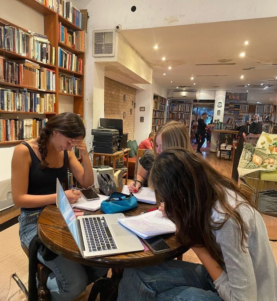 Three young women sit at a round table in a cozy, book-filled café, studying with notebooks, pens, and a laptop. Shelves of books line the walls, and other patrons can be seen in the background.