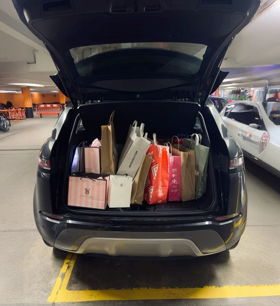 A black SUV parked in an indoor garage with its trunk open, filled with various colorful shopping bags and boxes. The surrounding area includes other parked cars and orange safety barriers.