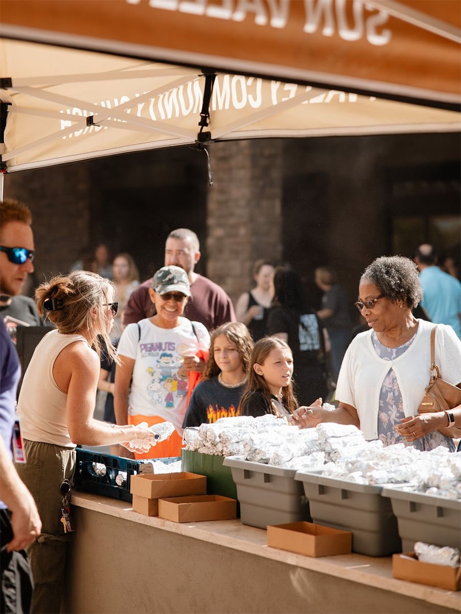 A woman hands food wrapped in foil to another woman under a tent at an outdoor event, with bins of wrapped food and people, including children, standing in line in the background.
