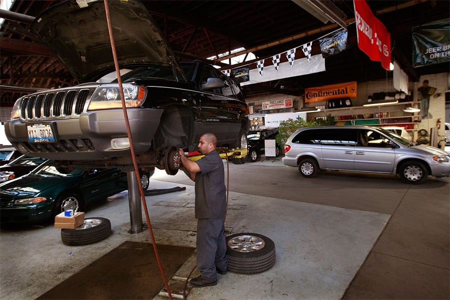 A mechanic works on the brake system of a raised SUV in an auto repair shop. Other vehicles and equipment are visible in the background, and banners hang from the ceiling.