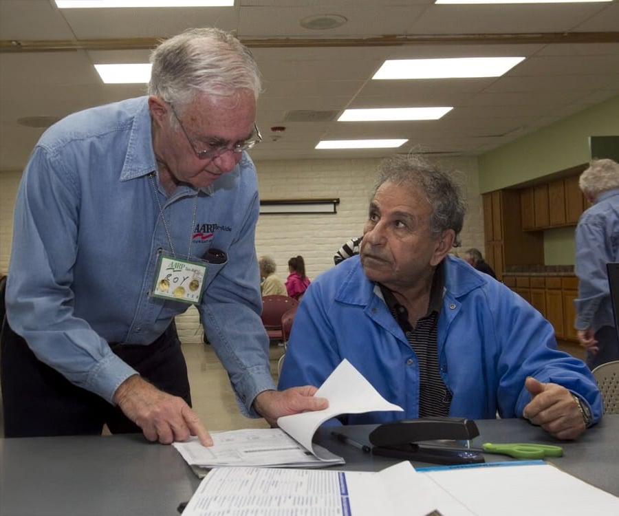 An older man standing and pointing at papers on a table helps another older man who is seated and looking up at him. Both men are wearing blue jackets in a community center or classroom setting.