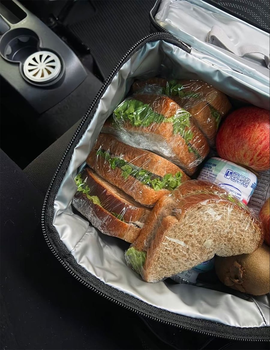 A lunch bag containing several wrapped sandwiches with lettuce, apples, a kiwi, and small yogurt containers, placed on a car seat next to a cup holder.
