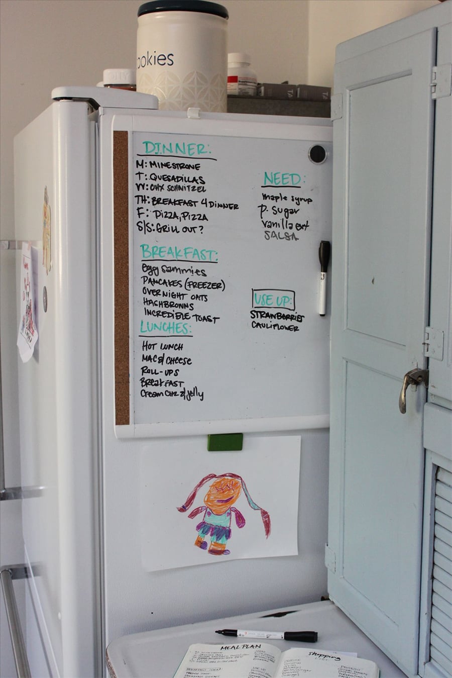 A whiteboard meal planner on a fridge lists dinner, breakfast, lunch, needed items, and foods to use up. A canister labeled "cookies" sits on top. A child's drawing is taped below the whiteboard. A cabinet is beside the fridge.
