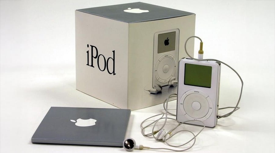 A first-generation Apple iPod with white earbuds is displayed next to its original box and a gray Apple-branded booklet, all set on a white surface.