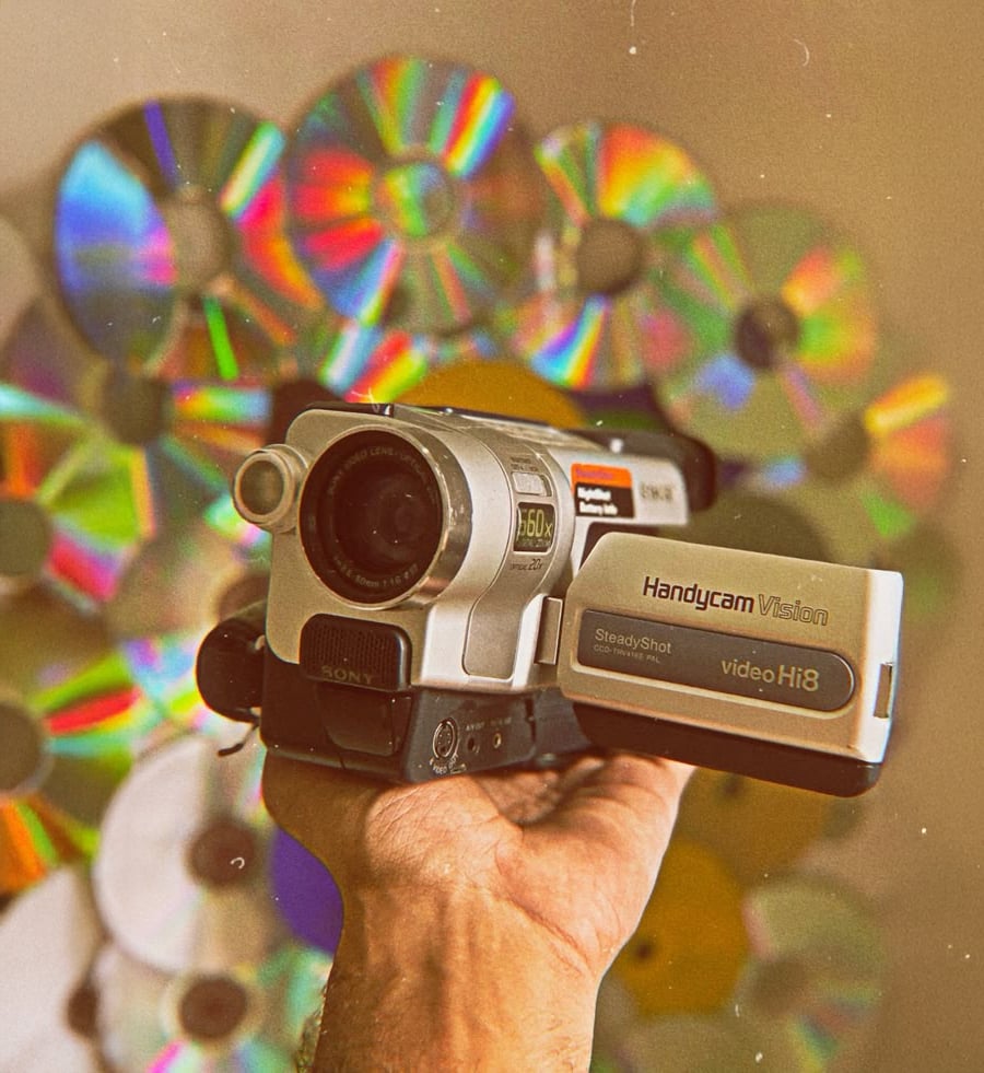 A hand holds a vintage Sony Handycam Vision camcorder, with a colorful, reflective backdrop made of CDs or DVDs arranged on a wall behind it.