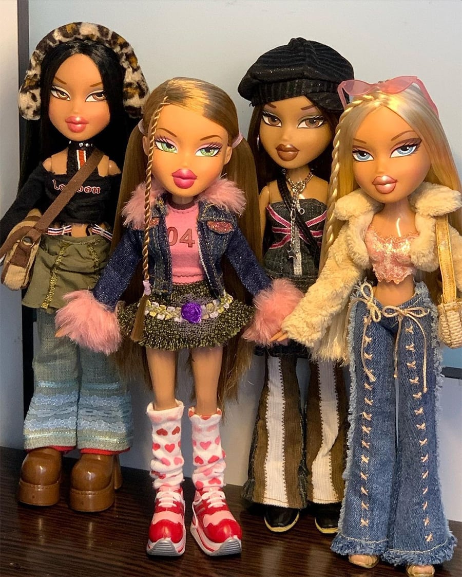 Four Bratz dolls stand side by side, each wearing colorful and trendy outfits with flared jeans, skirts, boots, and jackets, featuring long hair styled in different ways and bold makeup.