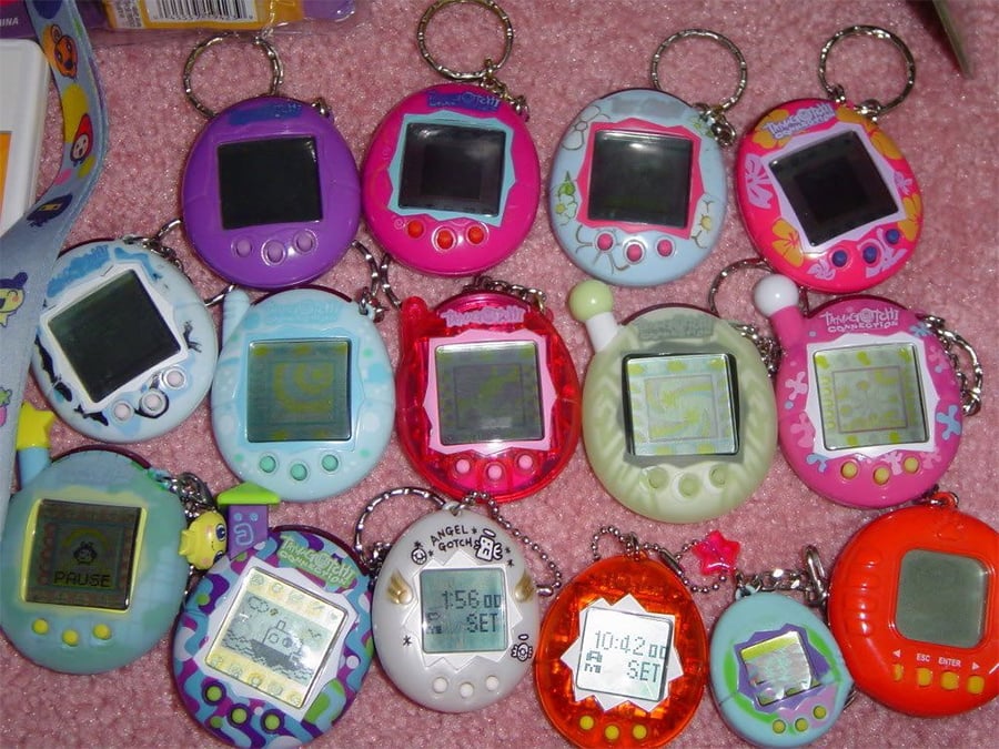 A collection of colorful Tamagotchi digital pets, including various shapes and designs, are arranged on a pink carpet. Each device has a small screen and attached keychain. Some screens display pixelated characters or menus.