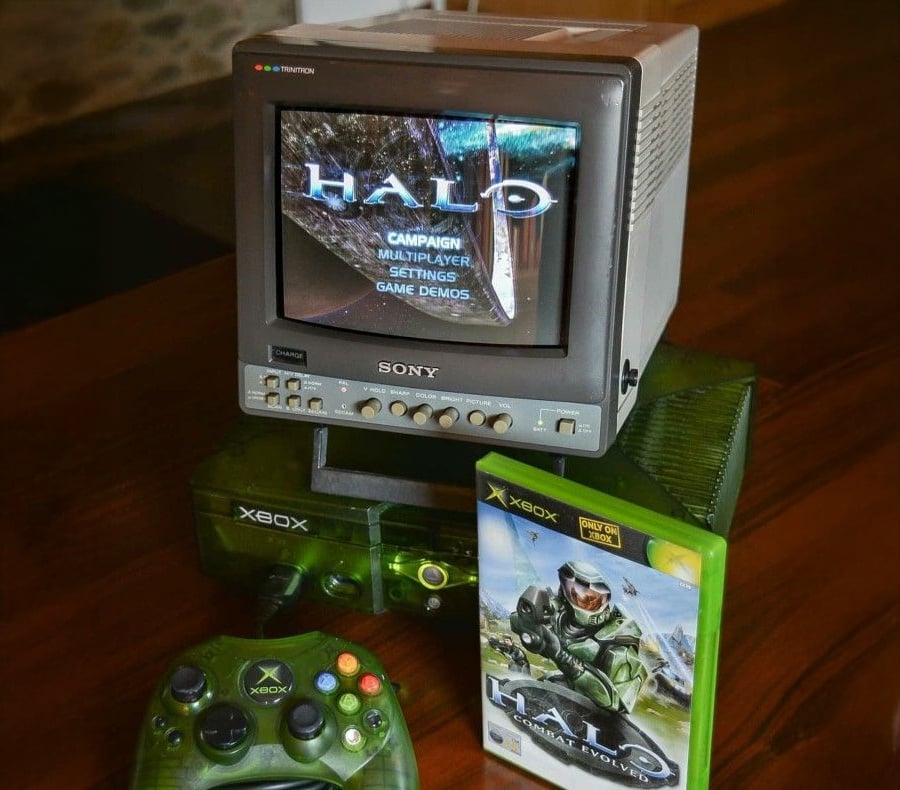 A classic Xbox console with a green controller and a "Halo: Combat Evolved" game case sits beside a small Sony CRT monitor displaying the Halo main menu screen.