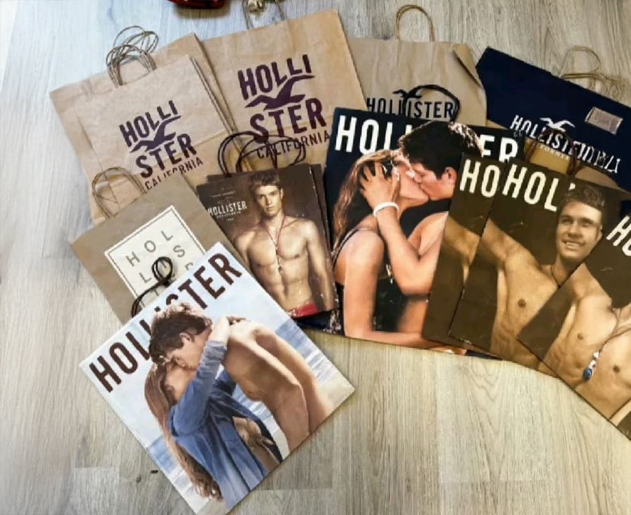 Several Hollister shopping bags are spread on a light wood floor. The bags feature images of models, including shirtless men and couples embracing, with the Hollister logo visible on each bag.