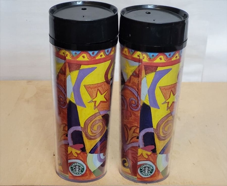 Two colorful Starbucks tumblers with abstract, vibrant designs featuring stars and swirls, standing side by side on a light-colored surface. Both have black lids and display the Starbucks logo at the bottom.