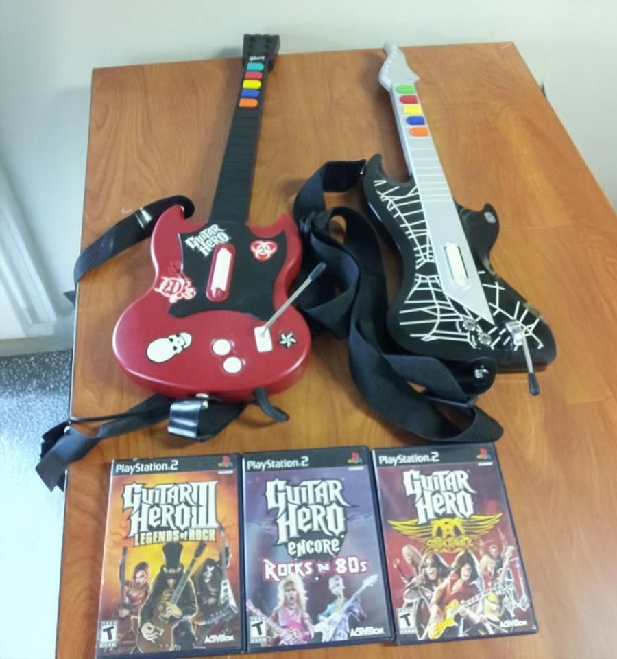 Two Guitar Hero controllers with themed designs and straps are on a wooden table, along with three PlayStation 2 Guitar Hero game cases: Legends of Rock, Rocks the 80s, and Guitar Hero II.