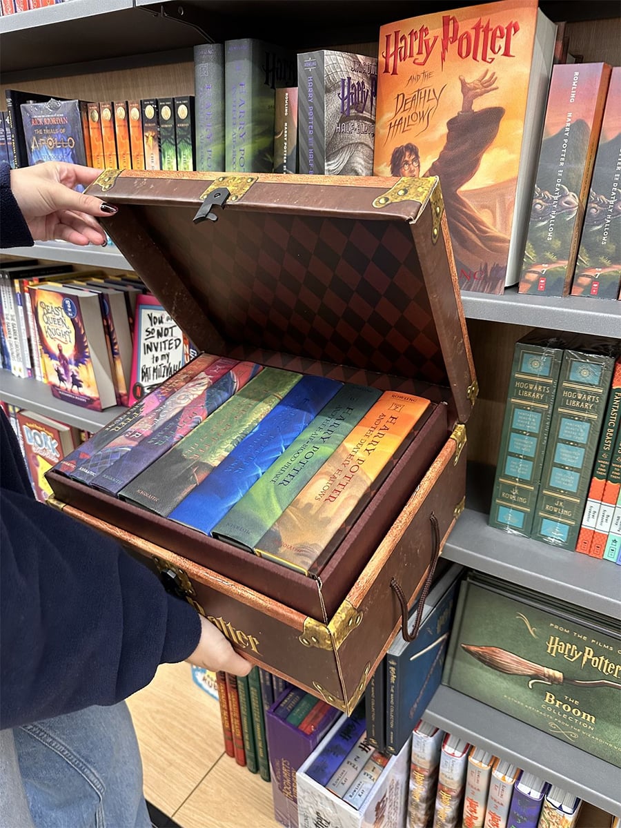 A person holds an open chest-shaped box set containing all seven Harry Potter books in colorful covers, displayed in a bookstore with Harry Potter books on the shelves.