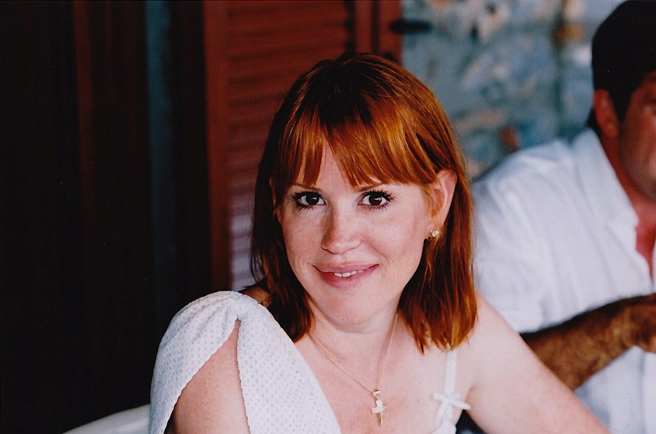 A woman with red hair, wearing a white top and a gold necklace, smiles at the camera. She is indoors, and a man partially visible sits beside her. The background is softly blurred.