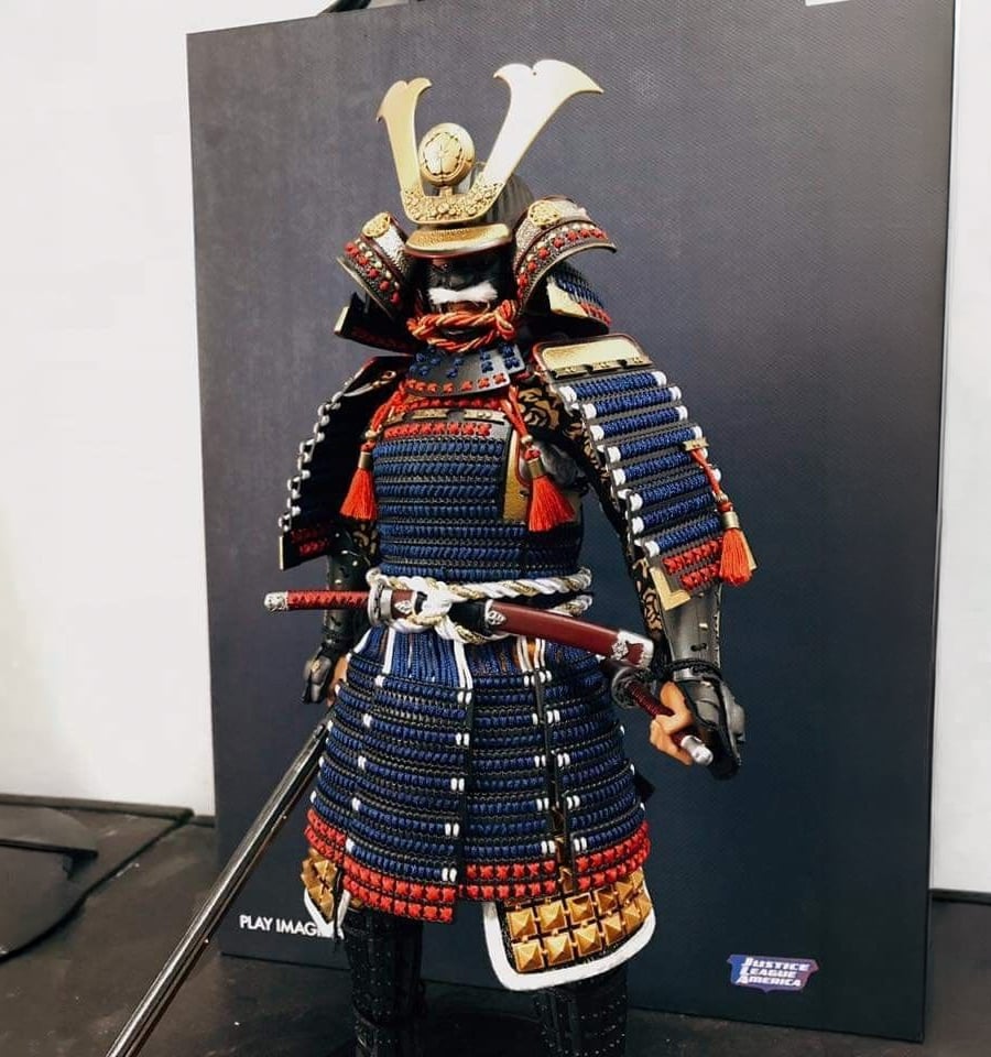 A detailed figurine of a samurai in blue and gold armor with red accents, holding a sword, stands in front of a dark rectangular display background.