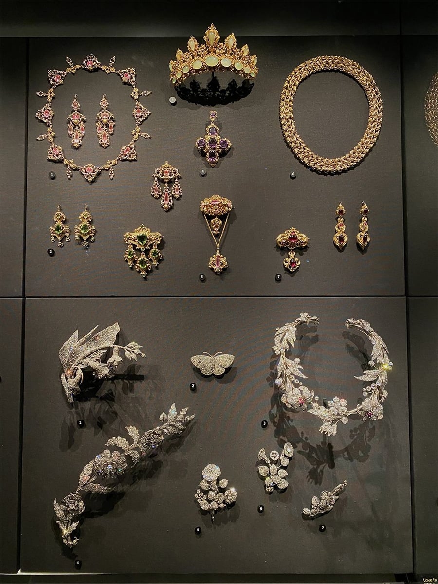 A museum display case with ornate jewelry sets including necklaces, earrings, brooches, tiaras, and hairpieces, featuring colorful gemstones and intricate designs, arranged on black velvet.