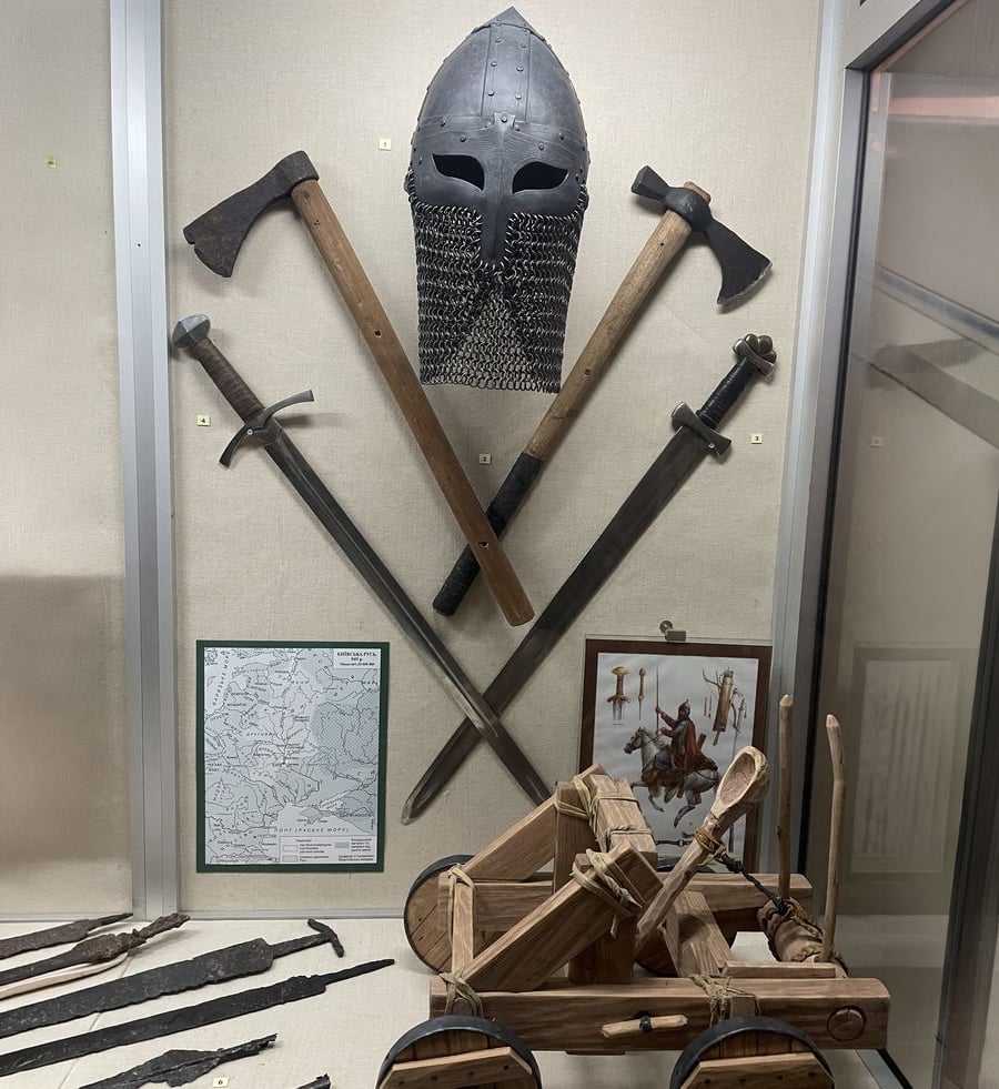 A museum display features a medieval helmet with chainmail, axes, swords, a map, a painting of a warrior, and a small wooden catapult model arranged neatly in a glass case.