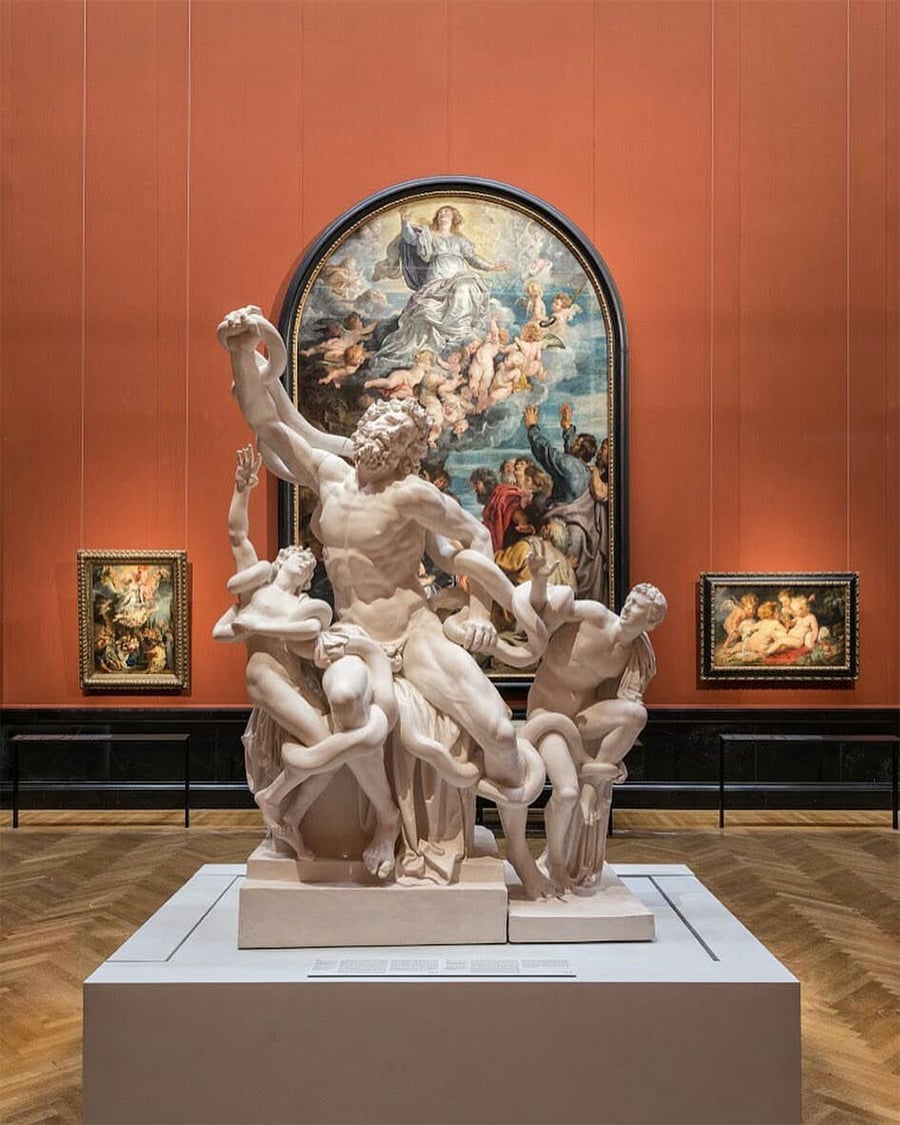 A marble sculpture of three men tangled with a serpent is displayed in a museum gallery with paintings on a red wall, including a large religious artwork in an arched frame directly behind the statue.
