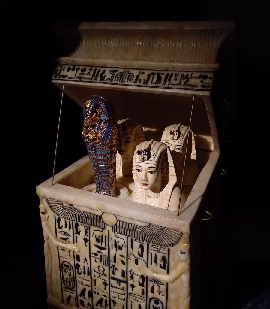 A decorative ancient Egyptian canopic chest with hieroglyphs, containing four intricately painted canopic jars with human and pharaoh faces, one adorned with blue and gold details, all displayed in a dimly lit setting.