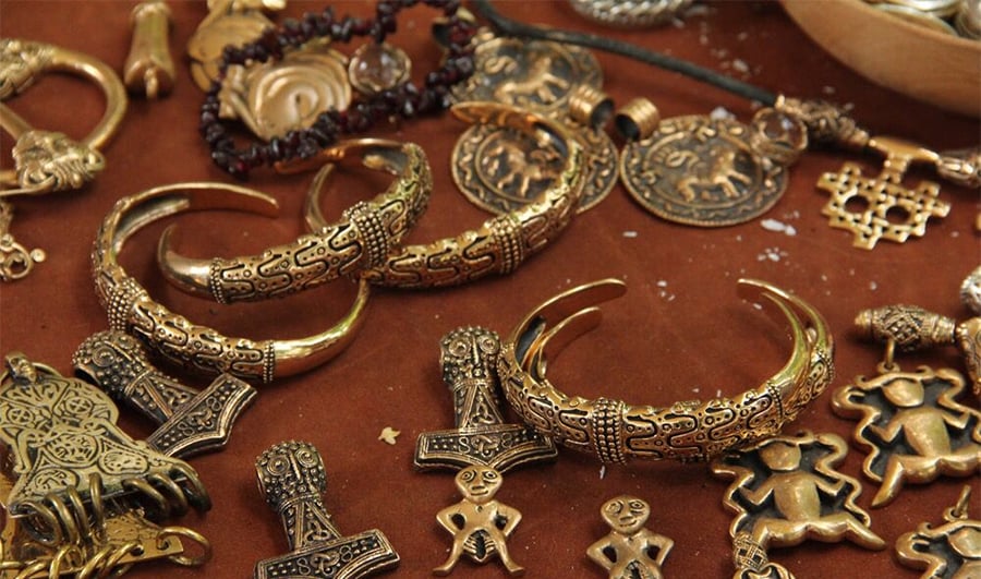 A collection of ornate, gold-colored Viking jewelry including bracelets, pendants, rings, and coins with intricate patterns and symbolic designs displayed on a brown surface.
