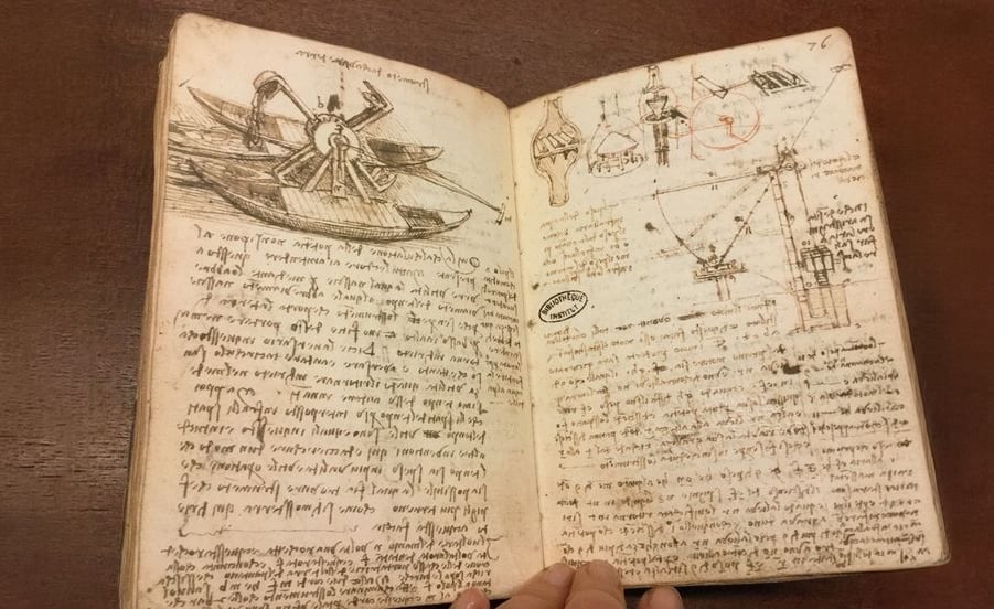 An open notebook with sketches of mechanical devices and handwritten notes in Italian, using mirror writing. The yellowed pages show detailed drawings and diagrams, resembling a historical inventor’s journal.