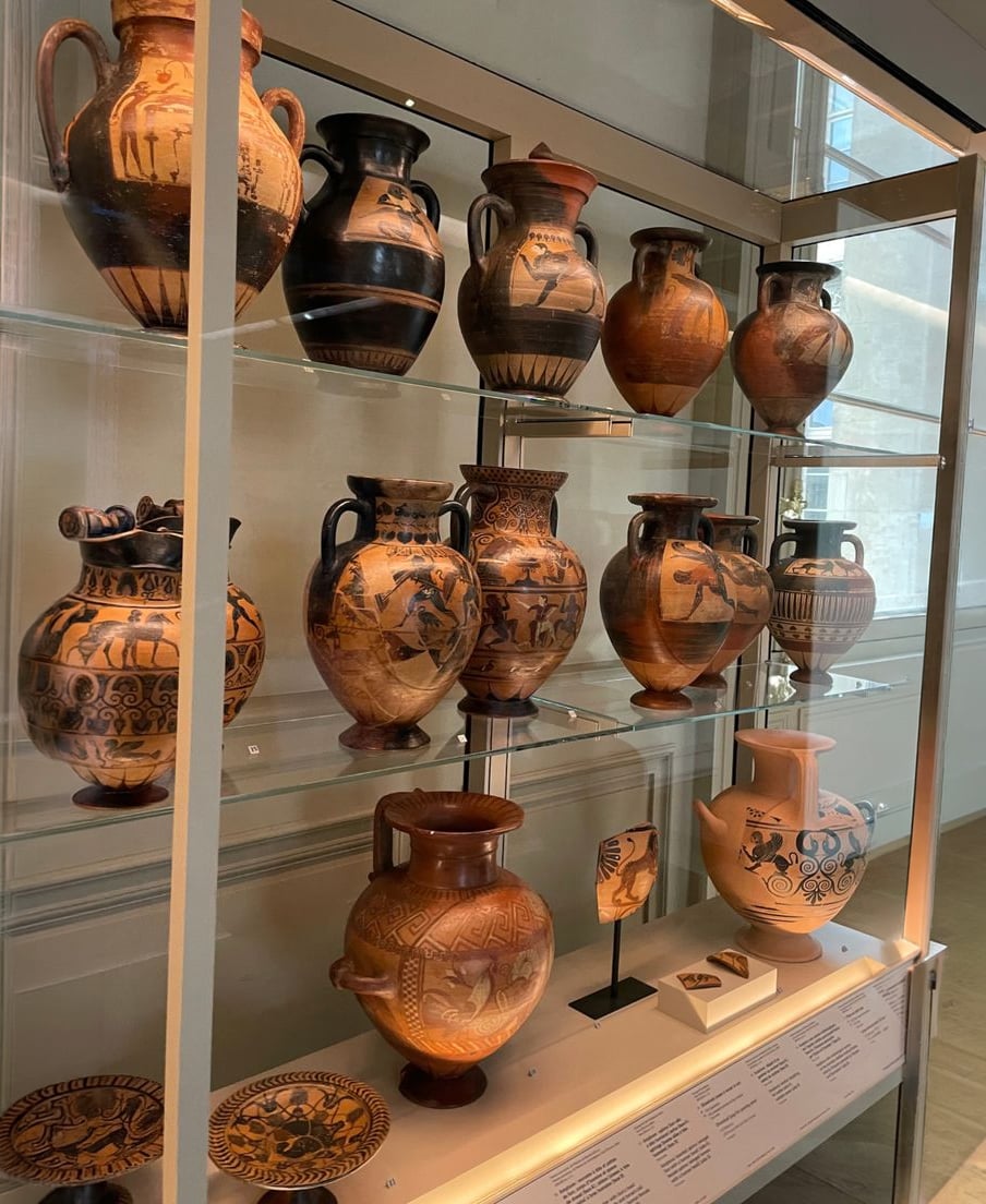 A display case containing ancient Greek pottery, including various painted vases and amphorae with intricate black and orange designs and figures, showcased on glass shelves in a well-lit museum room.