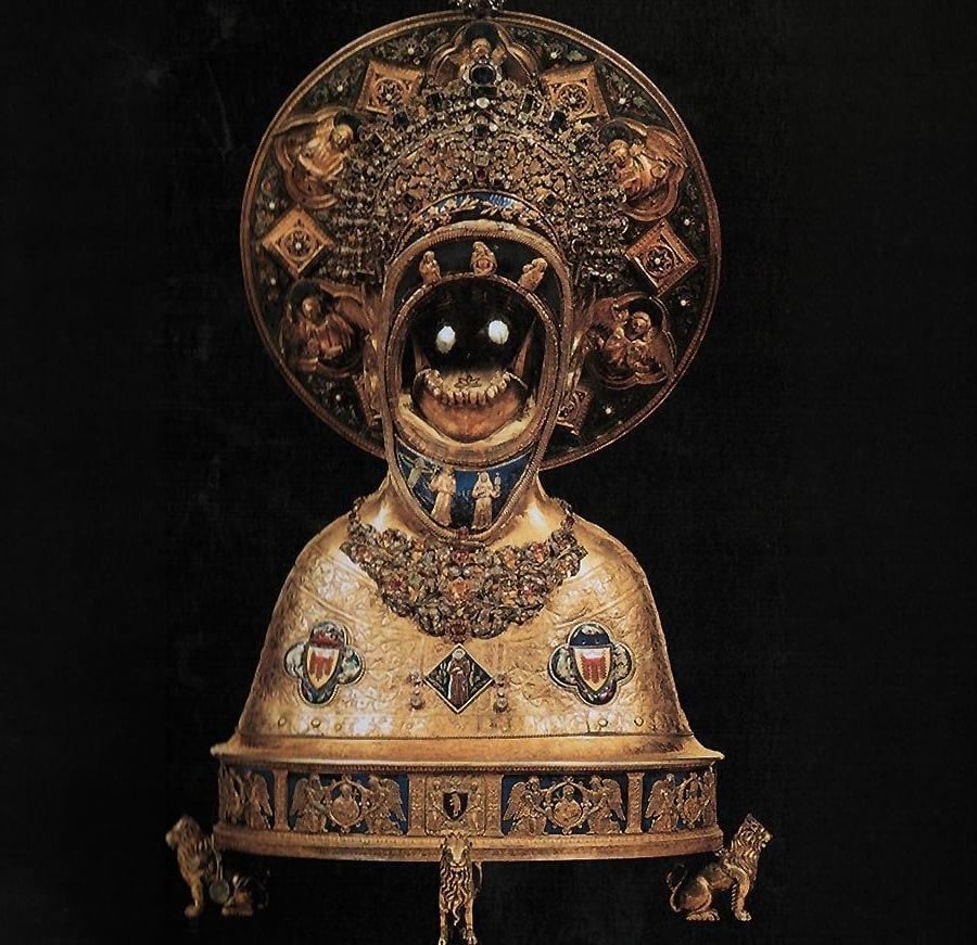 A golden, ornate medieval reliquary bust with gemstone eyes, a jeweled crown, and intricate religious decorations, set against a black background. The bust features heraldic shields and detailed metalwork.