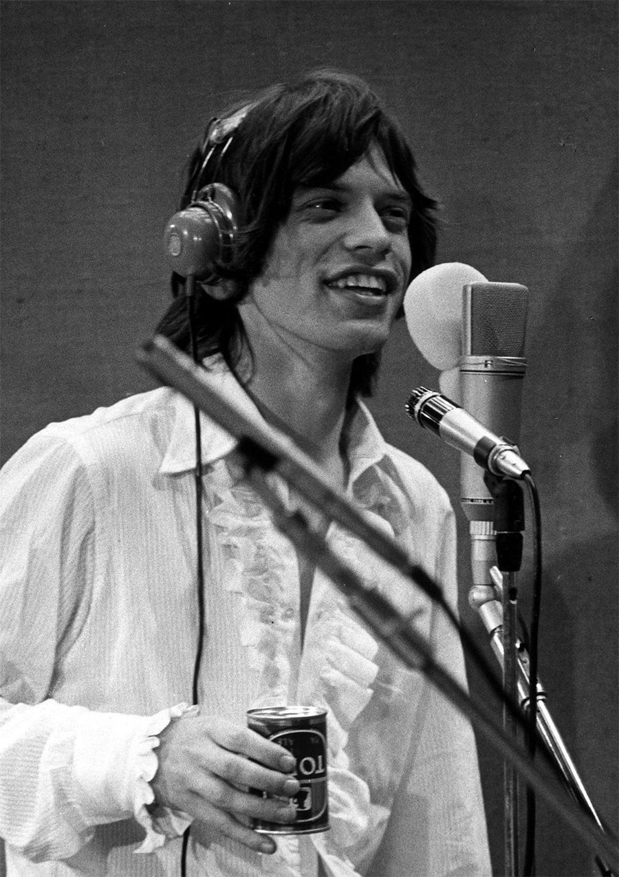 A young man with headphones and a ruffled shirt smiles while holding a soda can, standing in front of a studio microphone.