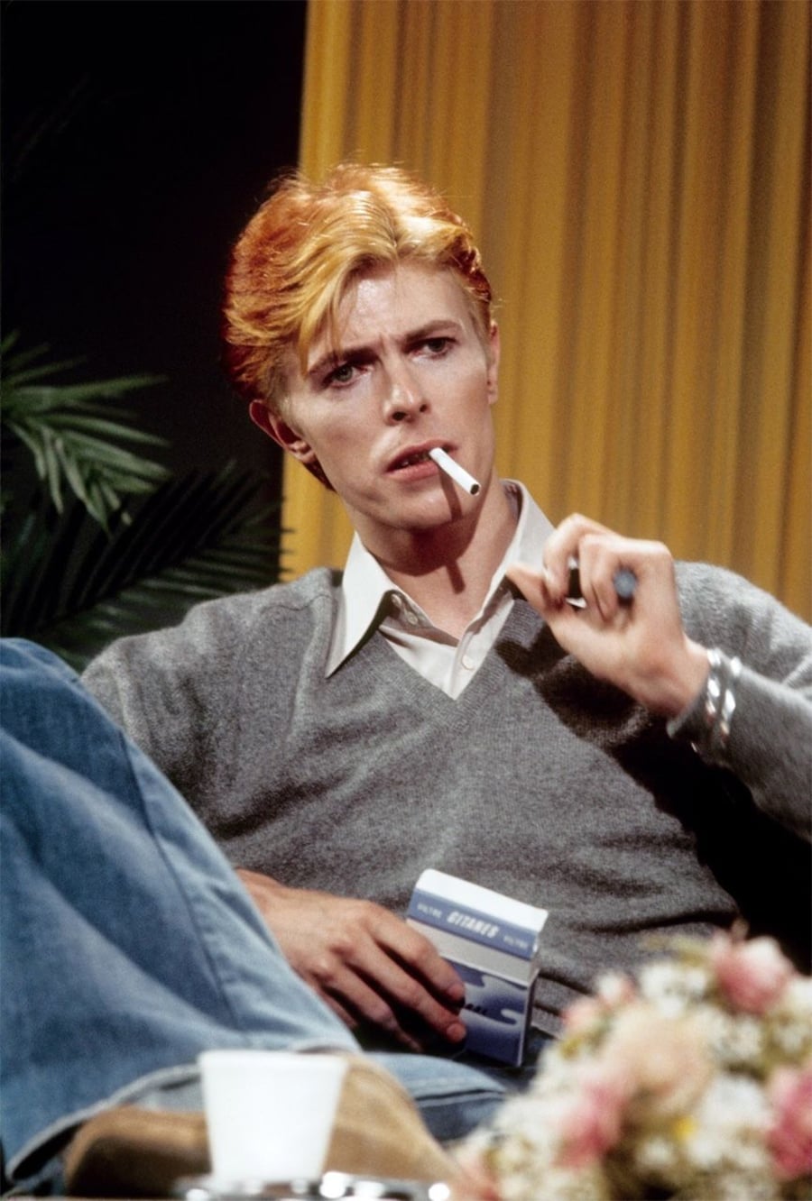 A person with orange-blonde hair sits casually with legs crossed, wearing a gray sweater over a white shirt and blue jeans, holding a cigarette and a pack of cigarettes, with a pensive expression. A yellow curtain is in the background.