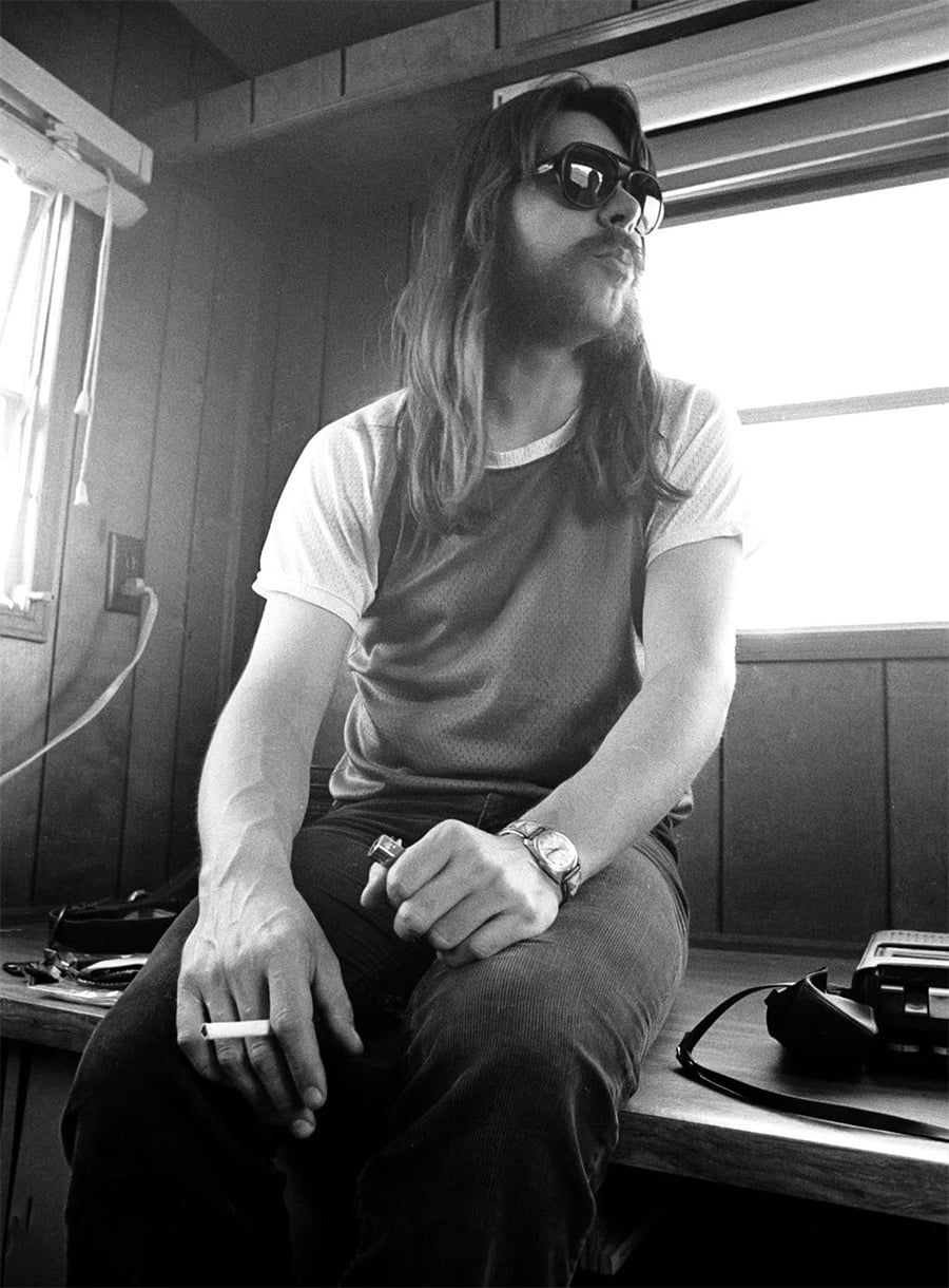 A man with long hair, a beard, and sunglasses sits on a desk by a window, holding a cigarette. He wears a short-sleeve shirt, jeans, and a watch, with headphones and an audio recorder nearby. The scene is in black and white.