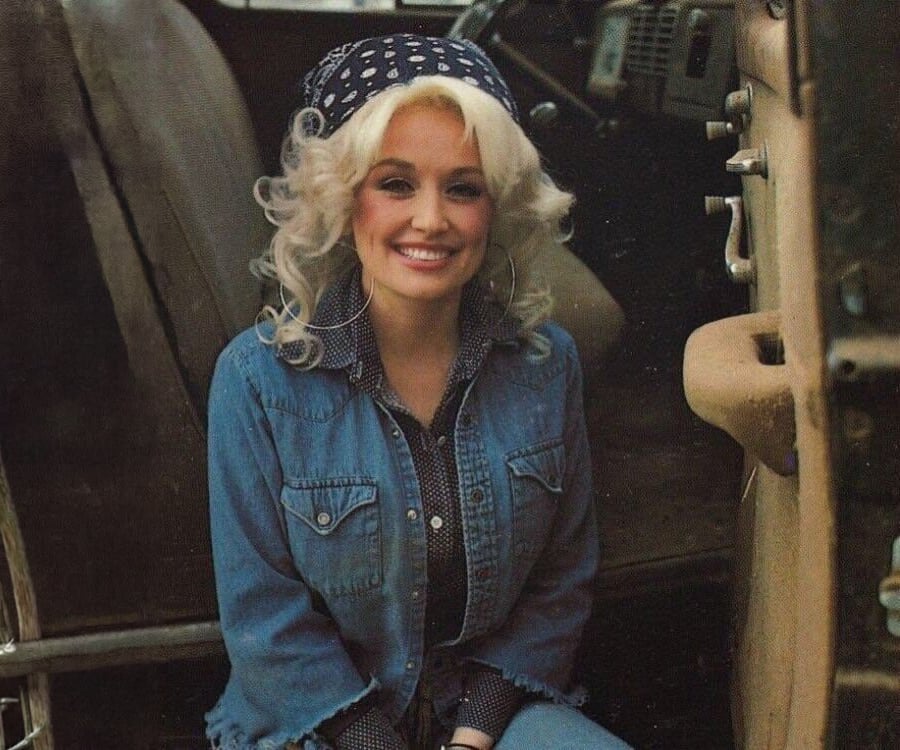 A woman with curly blonde hair, wearing a blue bandana, hoop earrings, a denim jacket, and jeans, smiles while seated inside an old vehicle with a rustic interior.