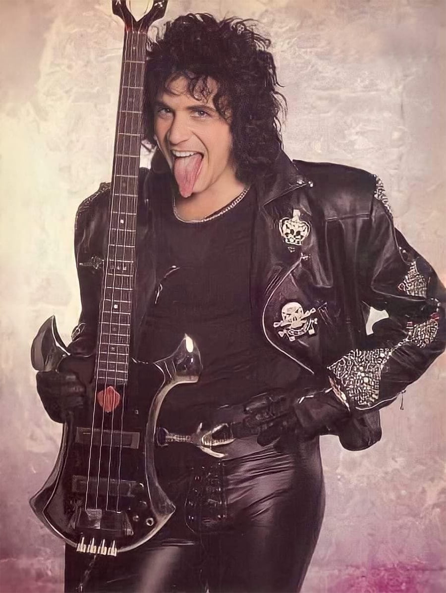 A person in a black leather jacket and pants stands with one hand on their hip and the other holding a uniquely shaped bass guitar. They have curly hair and are sticking out their tongue playfully. The background is light and neutral.