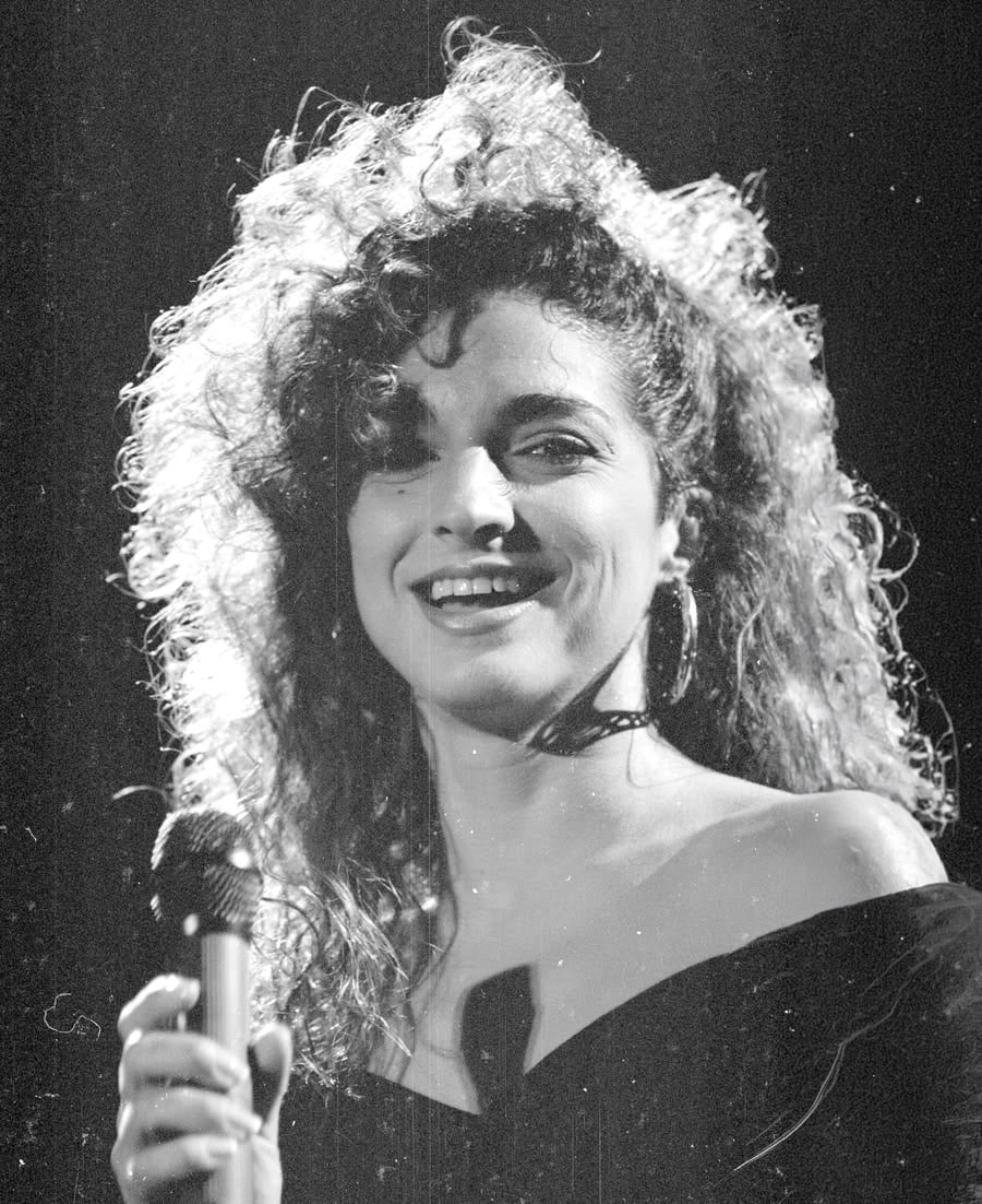 A woman with curly hair smiles while holding a microphone. She is wearing an off-the-shoulder top and hoop earrings, standing against a dark background with light shining on her face.