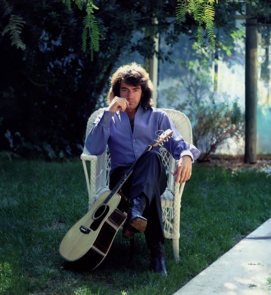 A man with long dark hair sits on a white wicker chair in a garden, wearing a purple shirt and black pants, with an acoustic guitar resting against his leg. Sunlight filters through the trees.