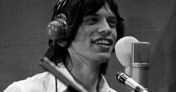A young man with long hair wearing headphones smiles while singing into a large microphone in a recording studio. The image is in black and white.