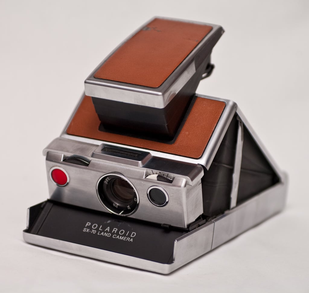 A vintage Polaroid SX-70 Land Camera with a silver body and brown leather accents is shown partially open, displaying its classic folding design against a plain white background.