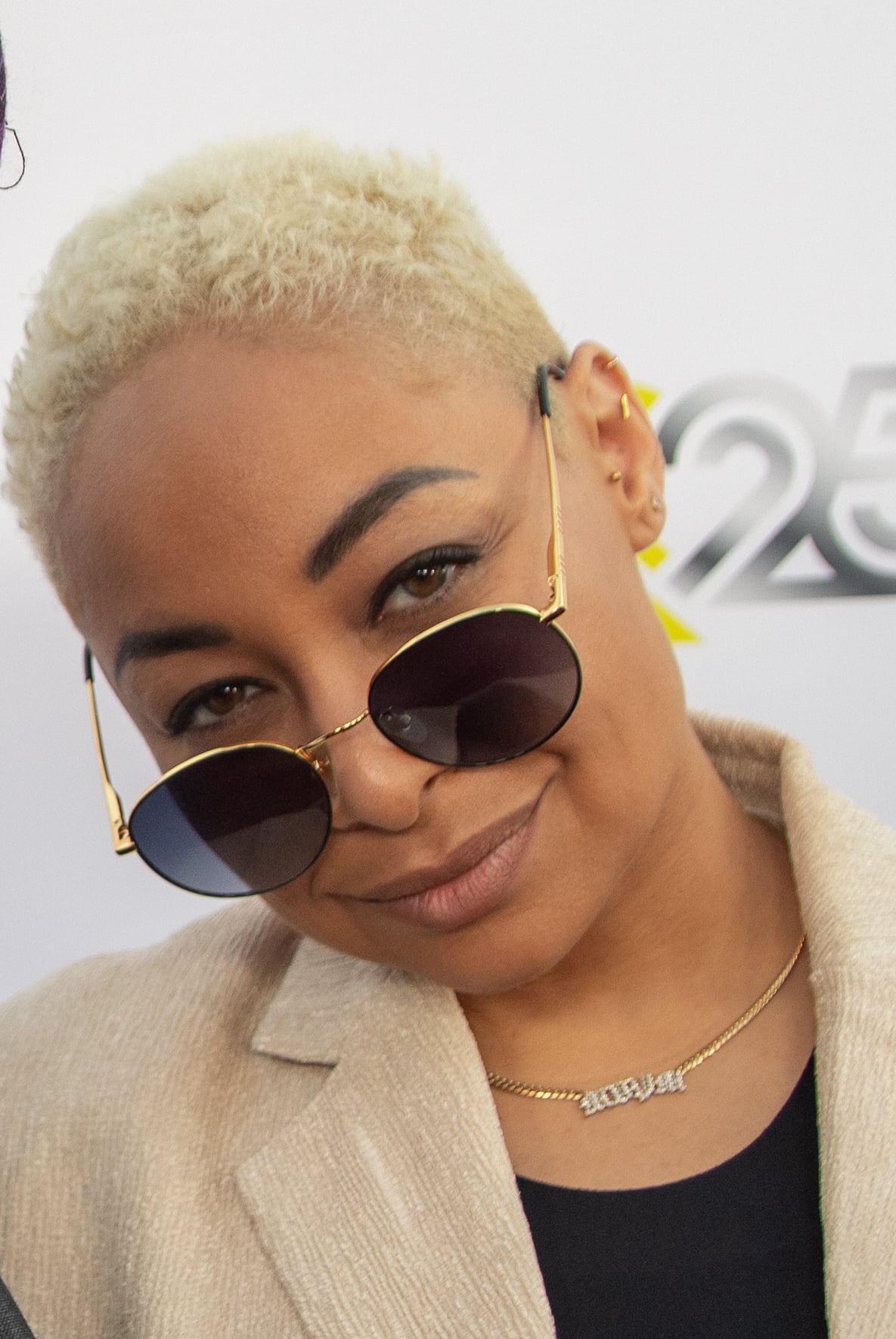 A person with short blond hair and dark sunglasses tilted down, wearing a beige blazer and a necklace, smiles slightly in front of a white background with "25" visible.