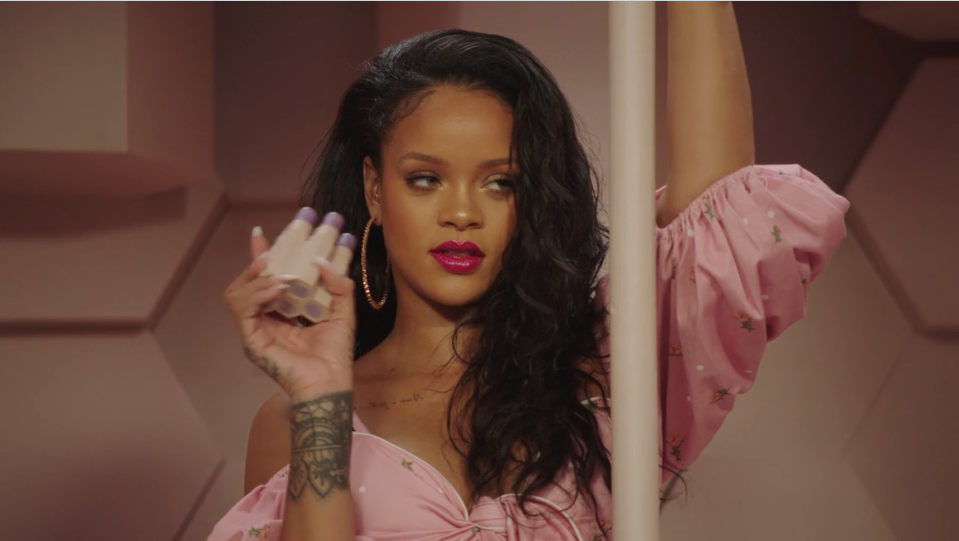 A woman with long black hair, wearing a pink off-shoulder top and hoop earrings, holds a makeup product and poses with one arm raised, standing against a geometric, peach-colored background.