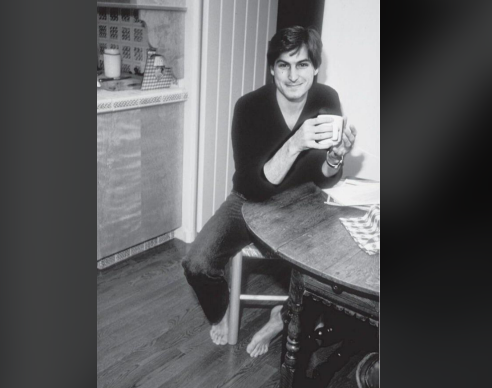 A man with dark hair, wearing a long-sleeve shirt and jeans, sits barefoot on a chair by a round wooden table, holding a mug and smiling in a cozy kitchen setting. The photo is in black and white.