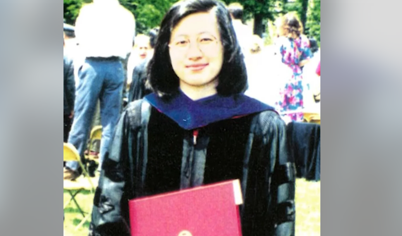 A graduate wearing a black gown and glasses holds a red diploma folder and smiles outdoors at a graduation ceremony, with people and trees in the background.