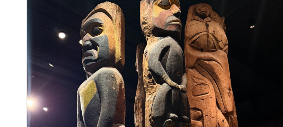 Three tall, carved wooden totem poles featuring human and animal figures are displayed indoors under spotlights, with intricate details and varying shades of brown and painted accents.
