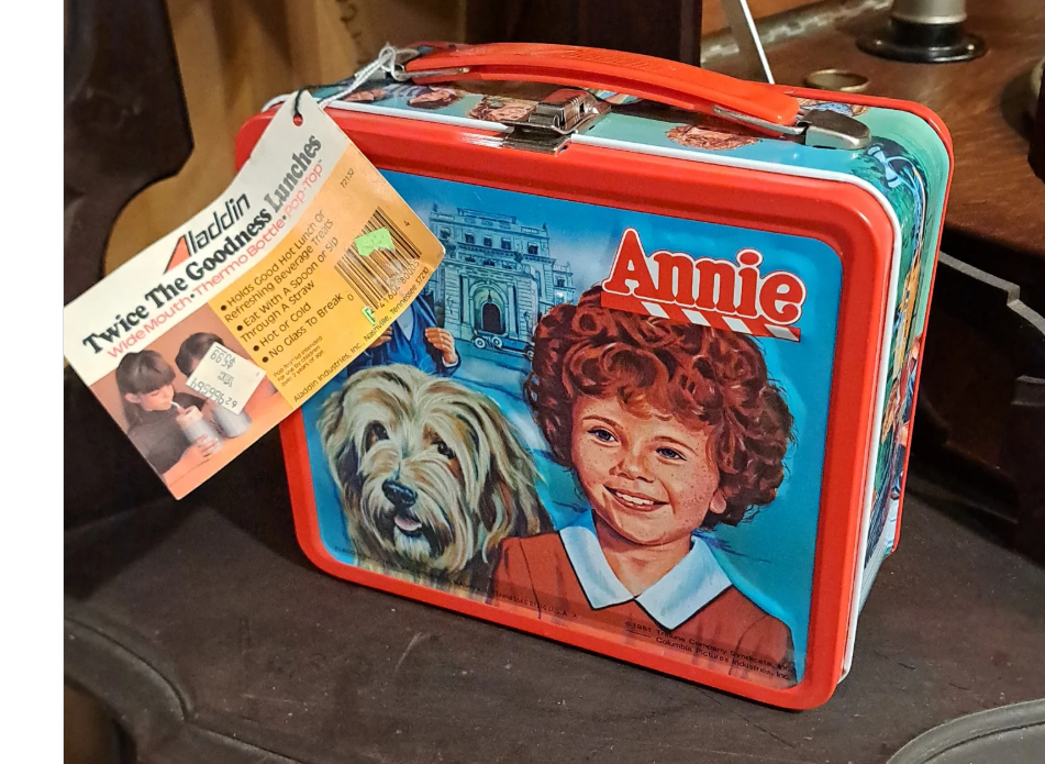 A vintage metal lunchbox featuring artwork of Annie, a curly-haired girl, and her dog Sandy, with a building and trees in the background. A product tag is attached to the handle.