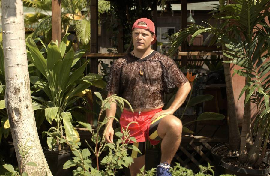 A person in a red cap, mesh shirt, and red shorts stands in a lush garden with one foot raised on a ledge, surrounded by various tropical green plants and wooden structures.